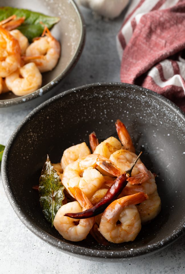 Gambas Al Ajillo (Spanish Garlic Shrimp) - A few pieces of shrimp in a black bowl topped with a red chili pepper
