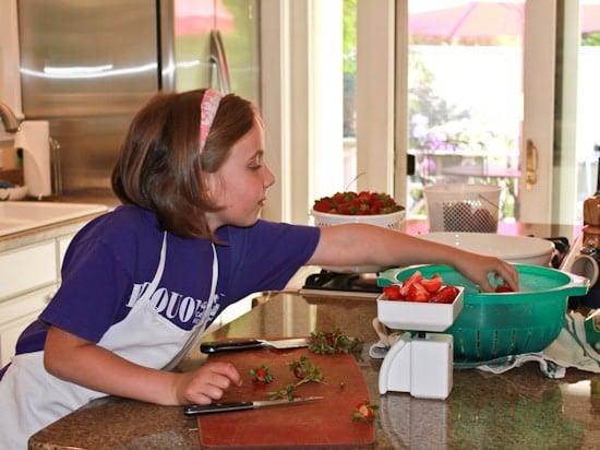 IMG_6056 A girl in a blue tee shirt, pink headband and white apron reaching across a kitchen countertop for more strawberries as she weighs them on a white set of old-fashioned scales.