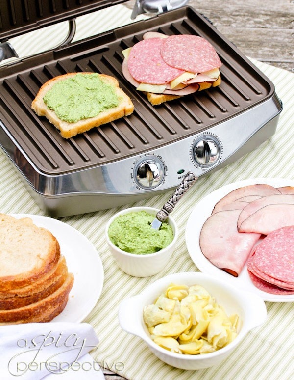 A panini maker is loaded with two slices of bread, one topped with creamy pesto and the other with salami and ham.
