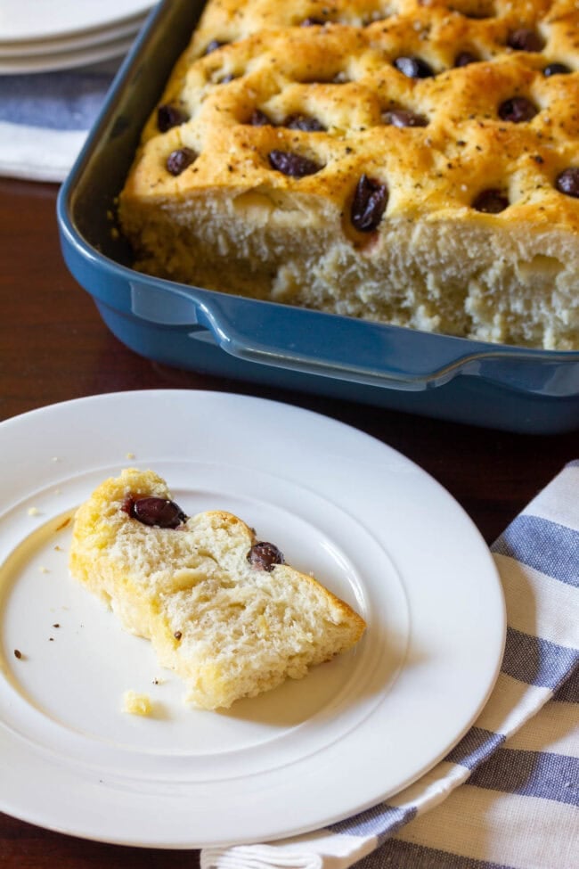 A loaf of focaccia is in a blue baking dish in the background. In the foreground is a white plate with a slice of fresh baked bread studded with olives.