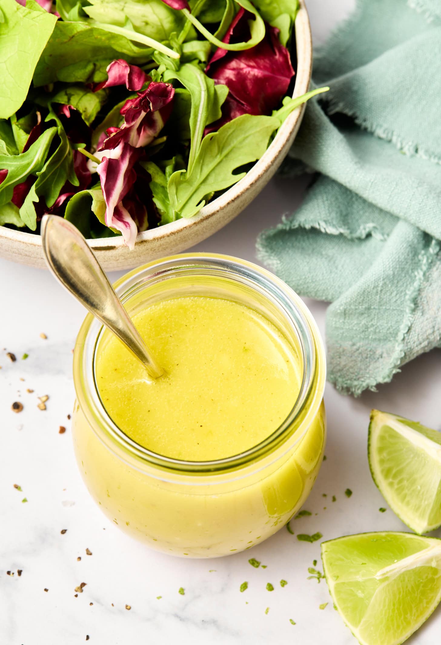 An overhead shot of a lime vinaigrette recipe in a glass jar, next to wedges of lime and a salad.