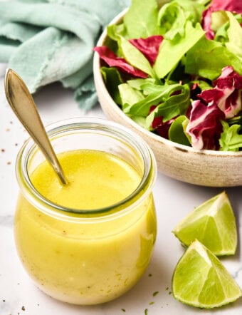 Easy lime vinaigrette in a glass jar with a spoon in it, next to a salad with fresh greens.