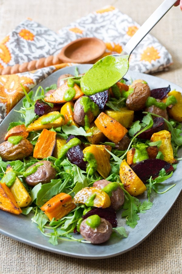 A hand drizzling homemade cilantro dressing over the roasted root salad on a serving platter.