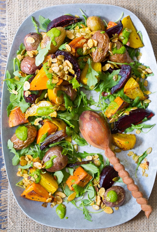 A top down view of roasted vegetable salad on a serving platter. A wooden spoon is resting on the platter where a few servings of the salad are missing.