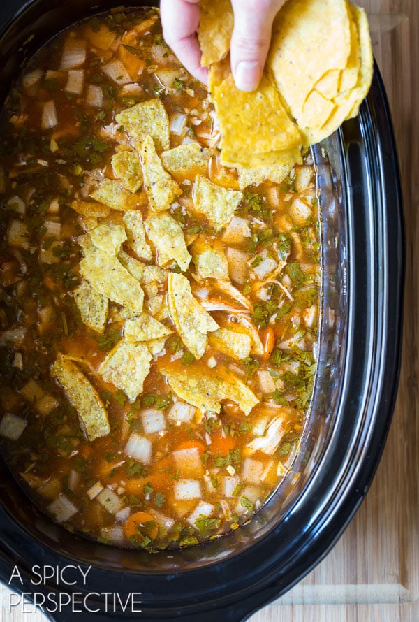 Hand adding crushed corn tortilla chips to the slow cooker of chicken soup.