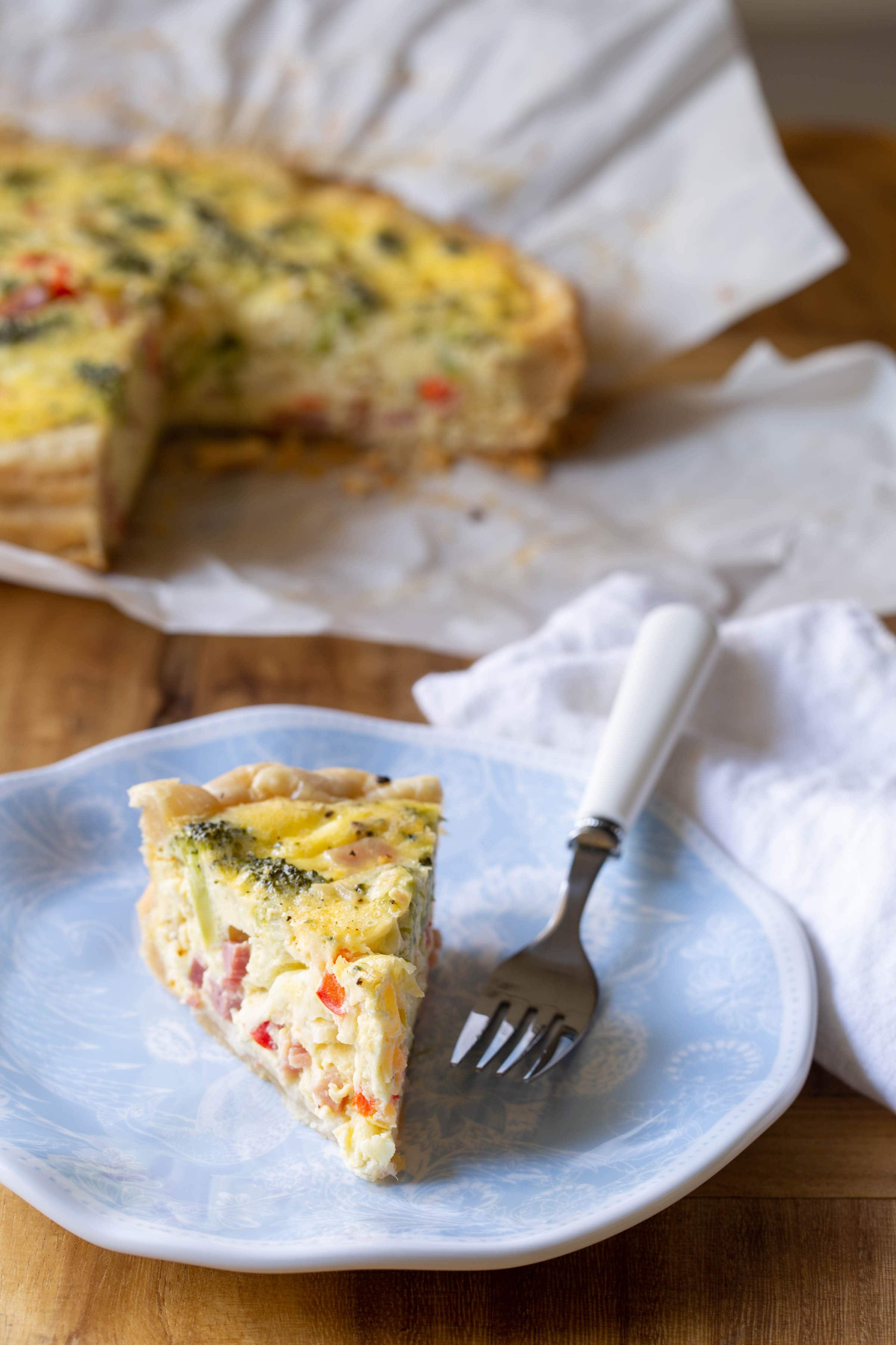 Single serving of breakfast quiche on a blue floral plate with a metal fork next to it. The full quiche is in the background on a piece of parchment paper.