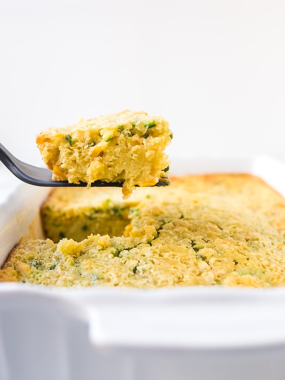 Side view of casserole dish and a spatula holding a piece of corn pudding 
