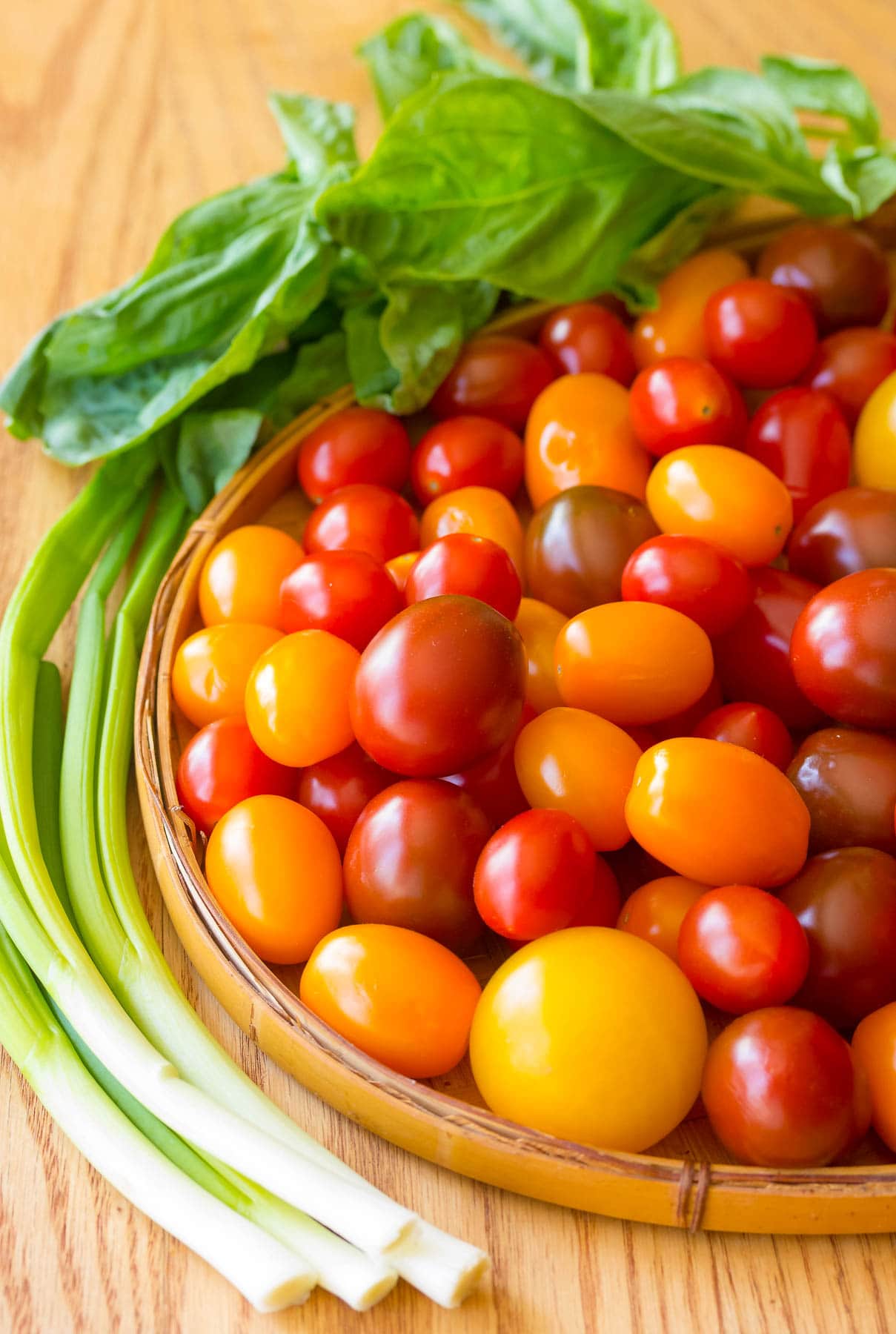 Basket of mixed cherry tomatoes with green onions next to it and picked basil in the background.