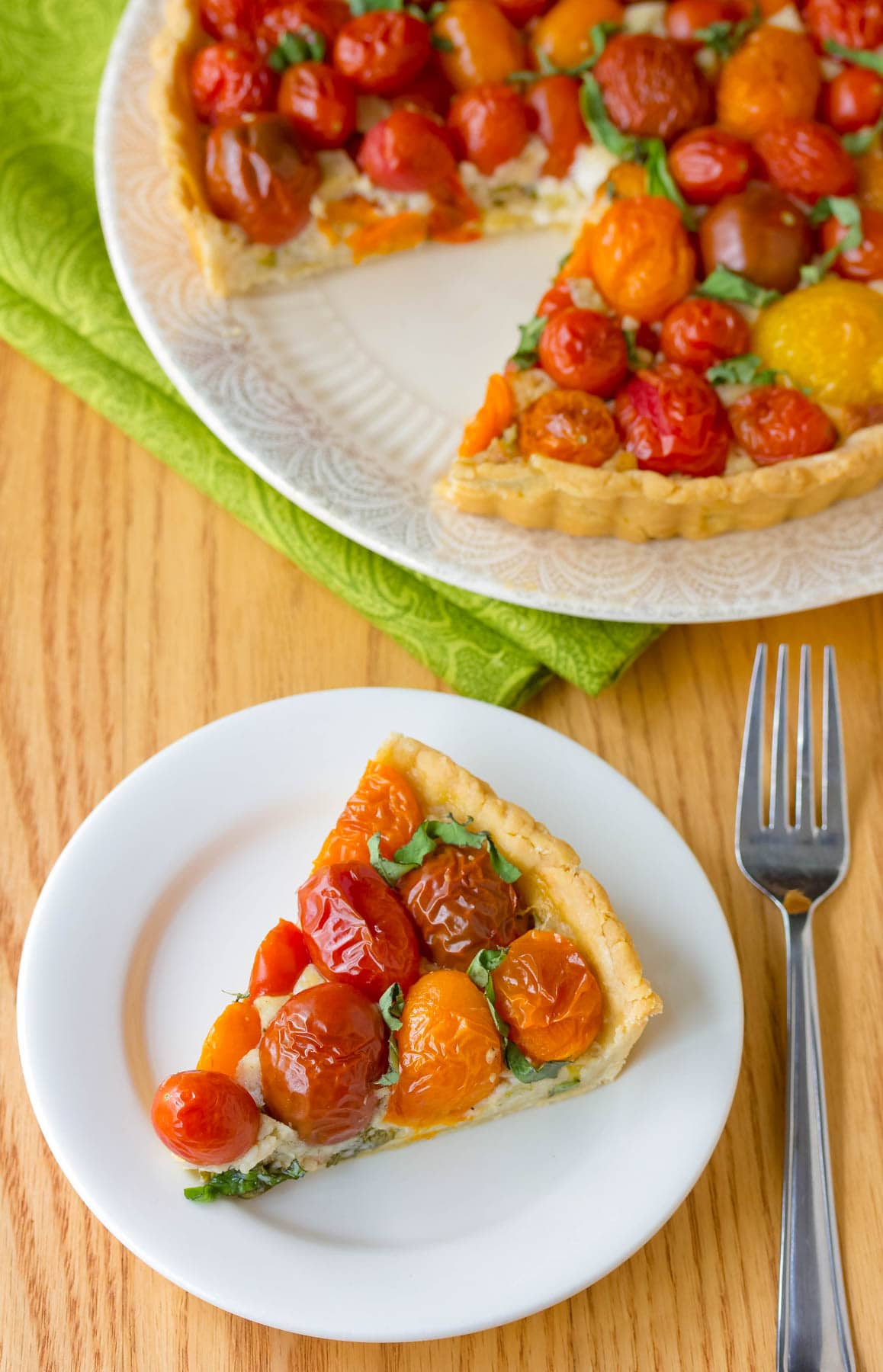 Slice of cherry tomato tart on a white plate with a fork next to it. The tart with a slice missing is on another white plate in the background.