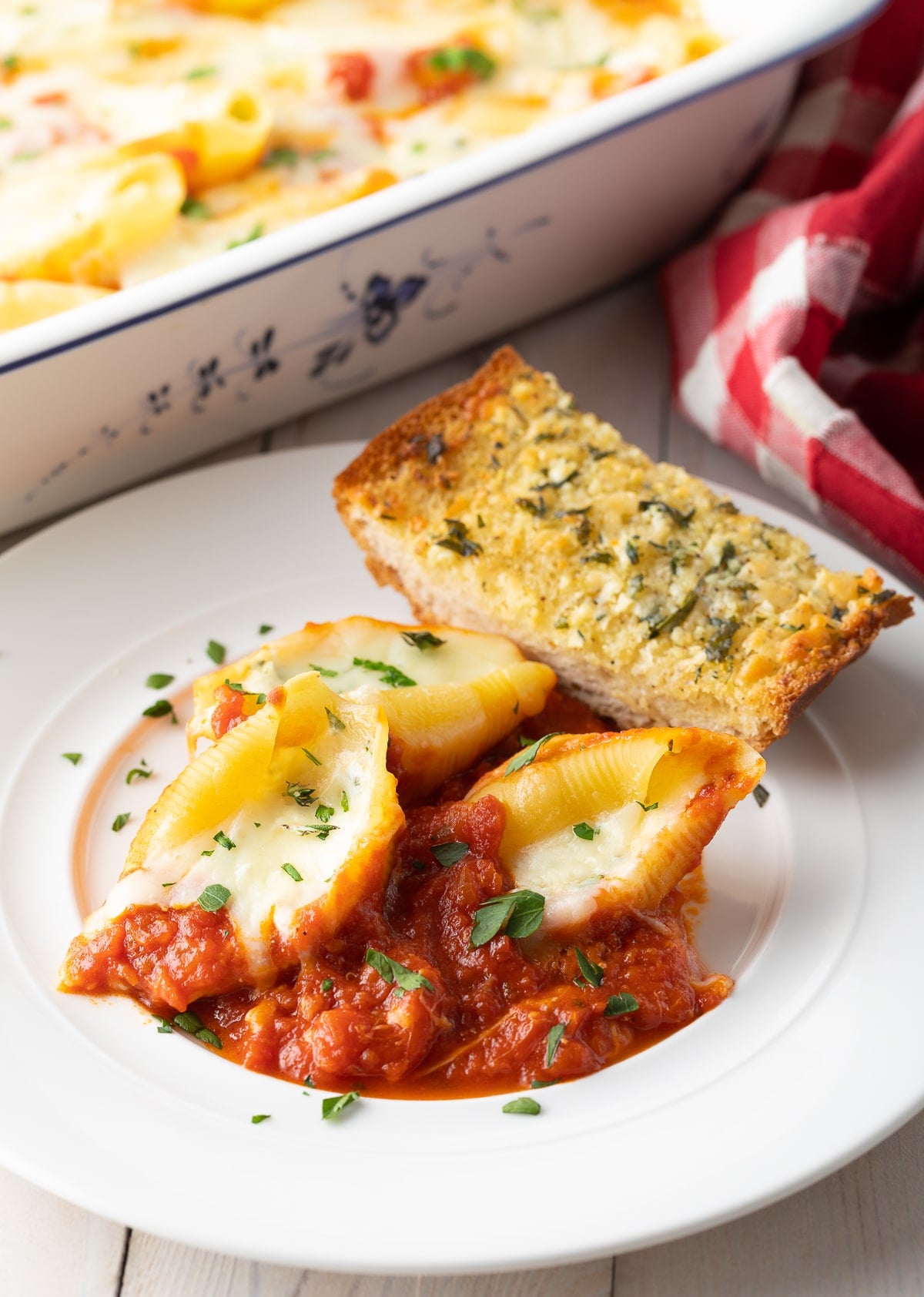 Ricotta stuffed shells in tomato sauce on a white plate with a piece of garlic bread on the side. The rest of the pasta bake is just visible in the background.