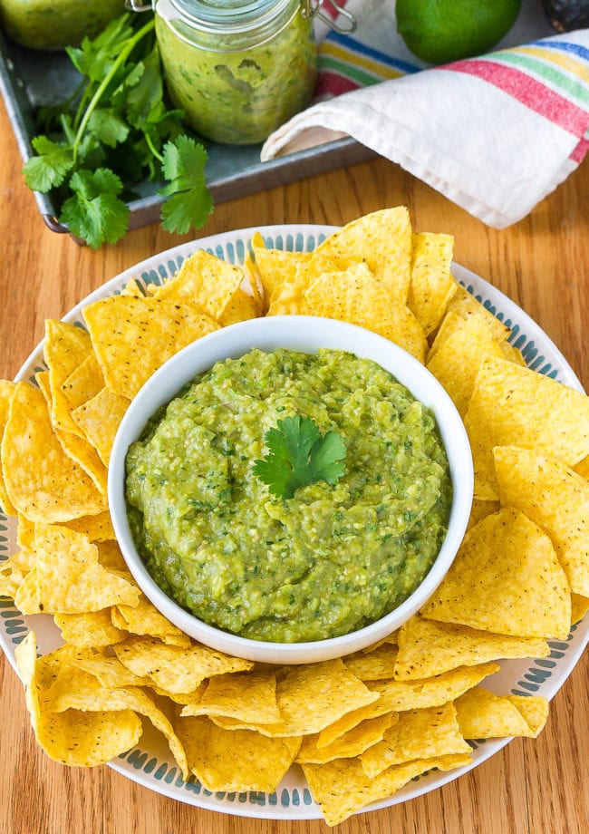 Small bowl full of tomatillo salsa verde on a plate, surrounded by tortilla chips. 