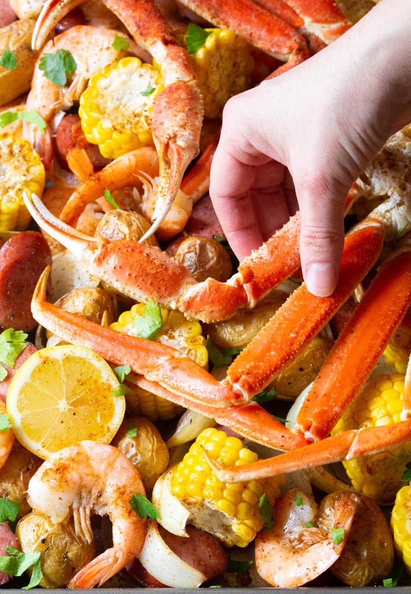 Hand picking up a long crab leg from low country seafood boil baked in the oven, with crab and shrimp nestled on a pan with cut ears of corn, whole baby potatoes, onions, and fresh lemon slices.