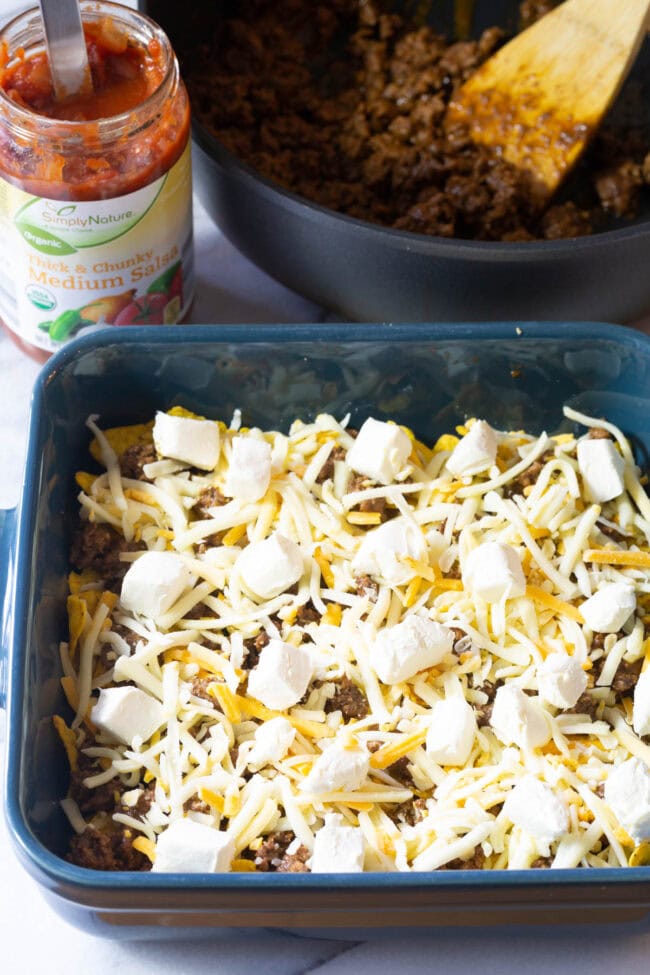 Doritos and minced meat topped with shredded cheese and cream cheese in baking dish.