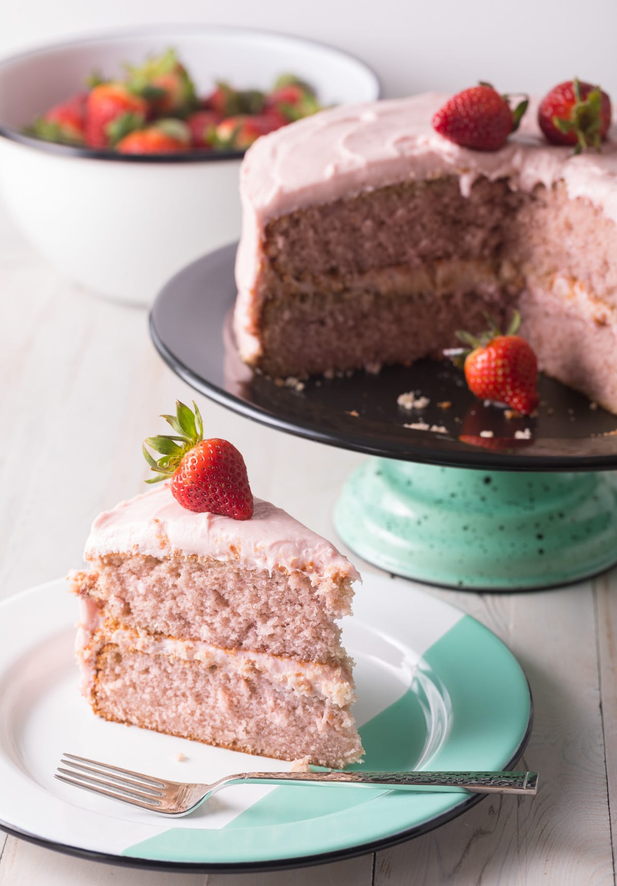 Whole fresh strawberry cake with pink frosting, topped with three fresh strawberries. A slice is removed and plated. 