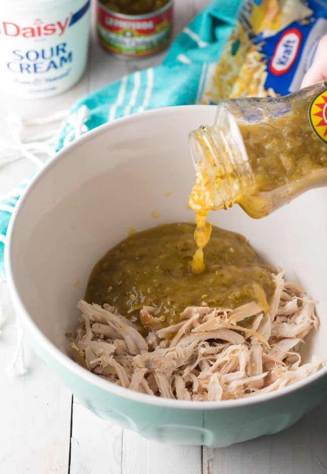 Pouring green salsa into a large bowl with shredded chicken.