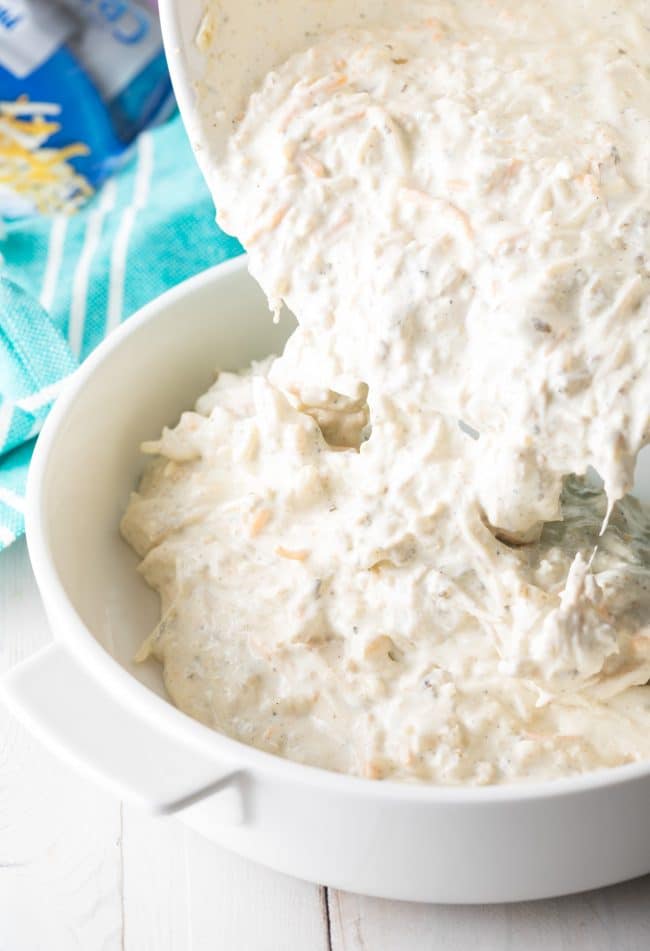 Pouring creamy sour cream and cheese dip into a prepared white round casserole dish.