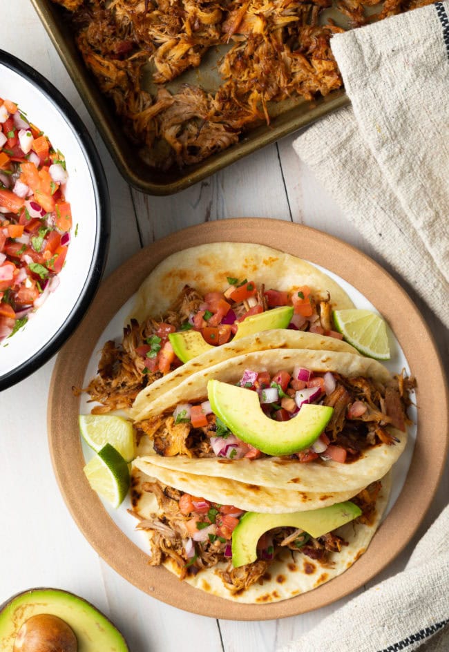 Overhead shot of plated slow cooker carnitas topped with fresh avocado slices and wedges of lime. 
