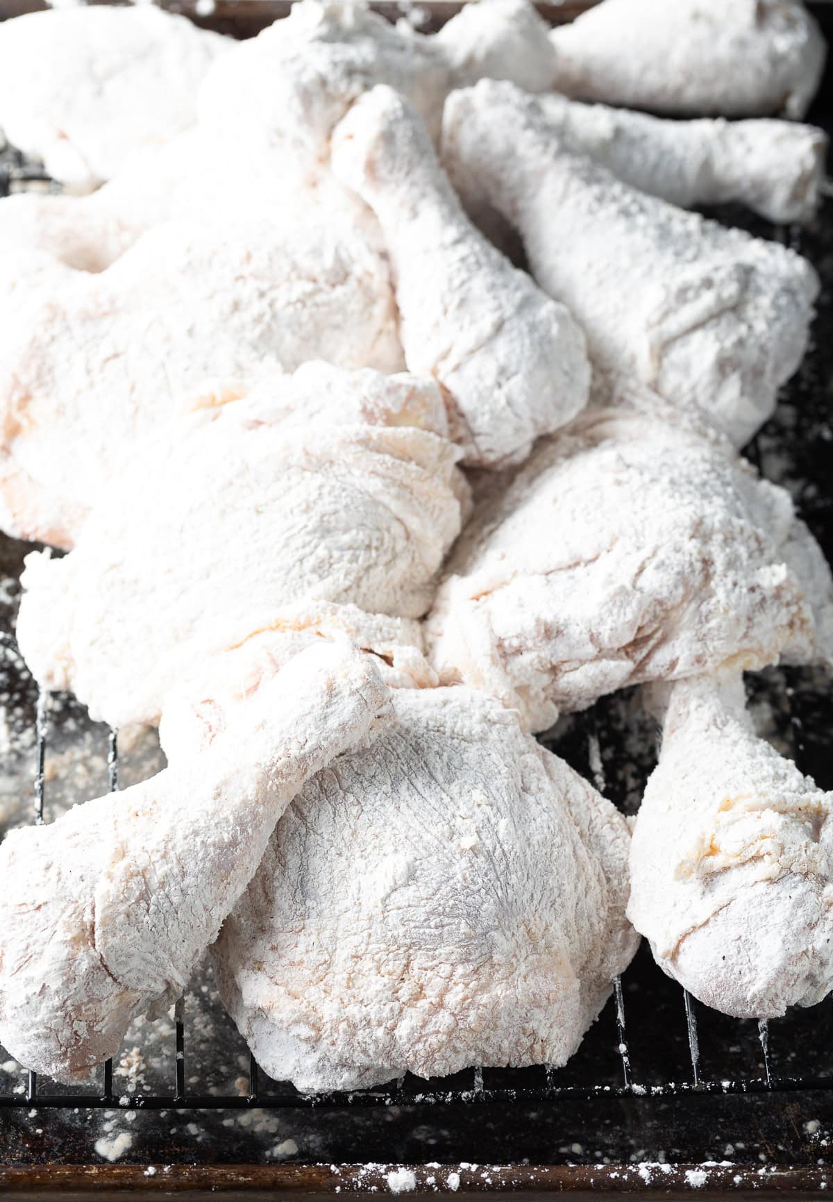 Several pieces of skin-on and bone-in chicken dredged in flour, laid out on a wire rack.