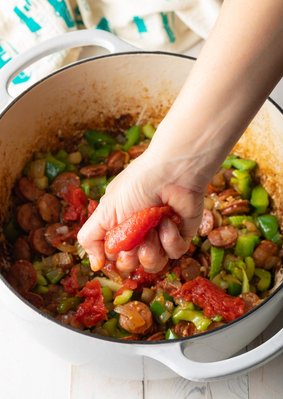 A hand squeezing a tomato over the pot of sausage and veggies.