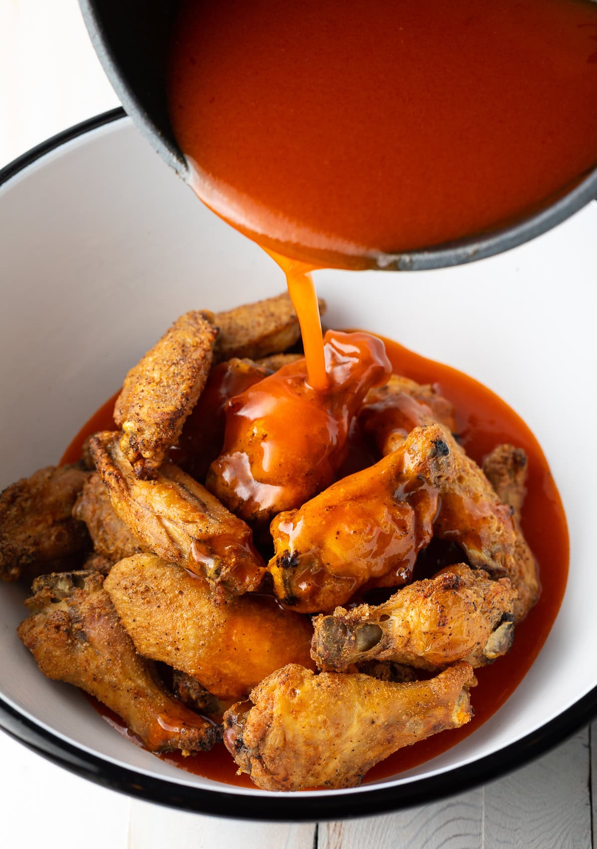 White mixing bowl with chicken wings, pouring buffalo sauce over the top.