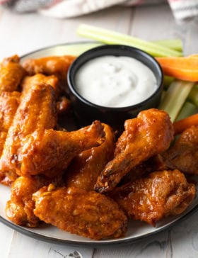 Plateful of baked buffalo chicken wings with black ramekin of ranch dressing, carrot sticks and celery sticks.