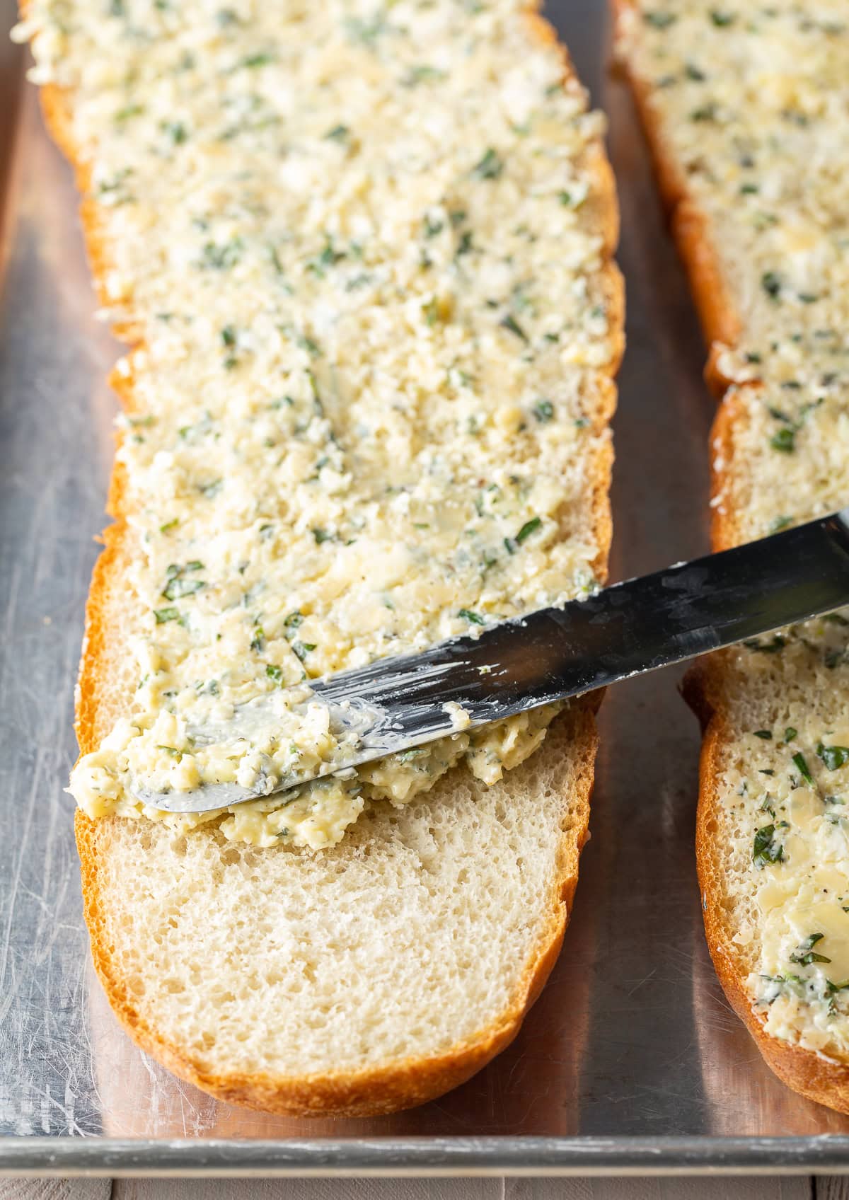 Knife smearing garlic butter herb mixture onto bread.