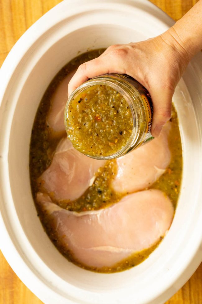 Slow cooker full of chicken breasts with some salsa verde being poured in 
