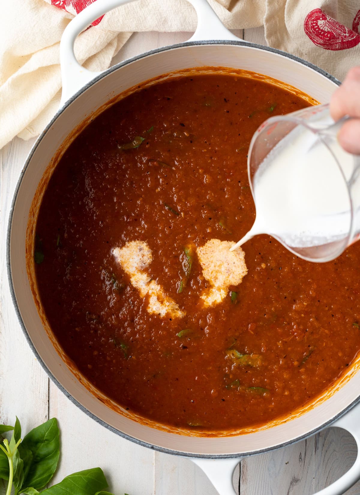 Tomato soup in a pot with a hand pouring heavy cream into the soup. 