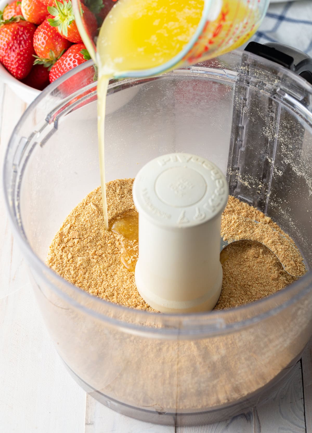 Butter being poured into food processor with finely pulsed graham crackers. 