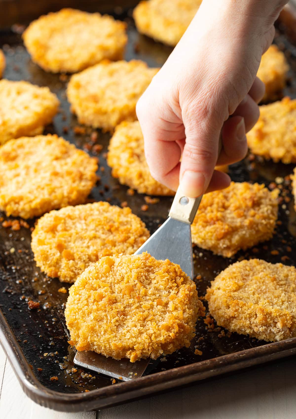 Hand using a spatula to pick up a crispy fried green tomato from the baking sheet.