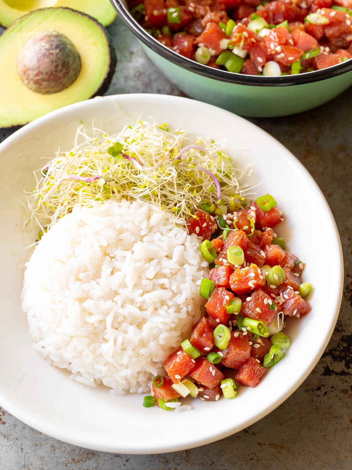Half-assembled poke bowl with rice, ahi tuna and spouts. There is the rest of the ahi tuna and half and avocado in the background.