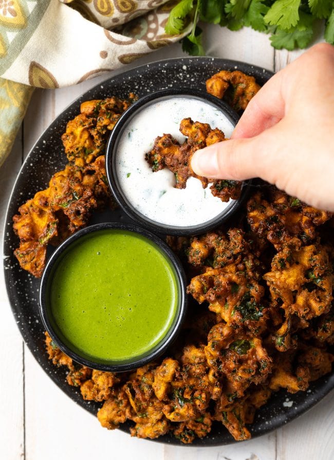 Top down view of a hand dipping a piece of crispy vegetable pakora into a ramekin of white sauce.