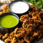 Plate with kale vegetable pakora, with a ramekin of white sauce and ramekin of green sauce.