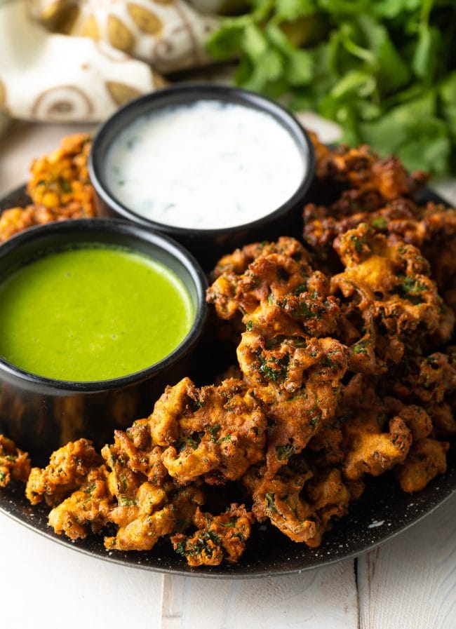 Plate with kale vegetable pakora, with a ramekin of white sauce and ramekin of green sauce.
