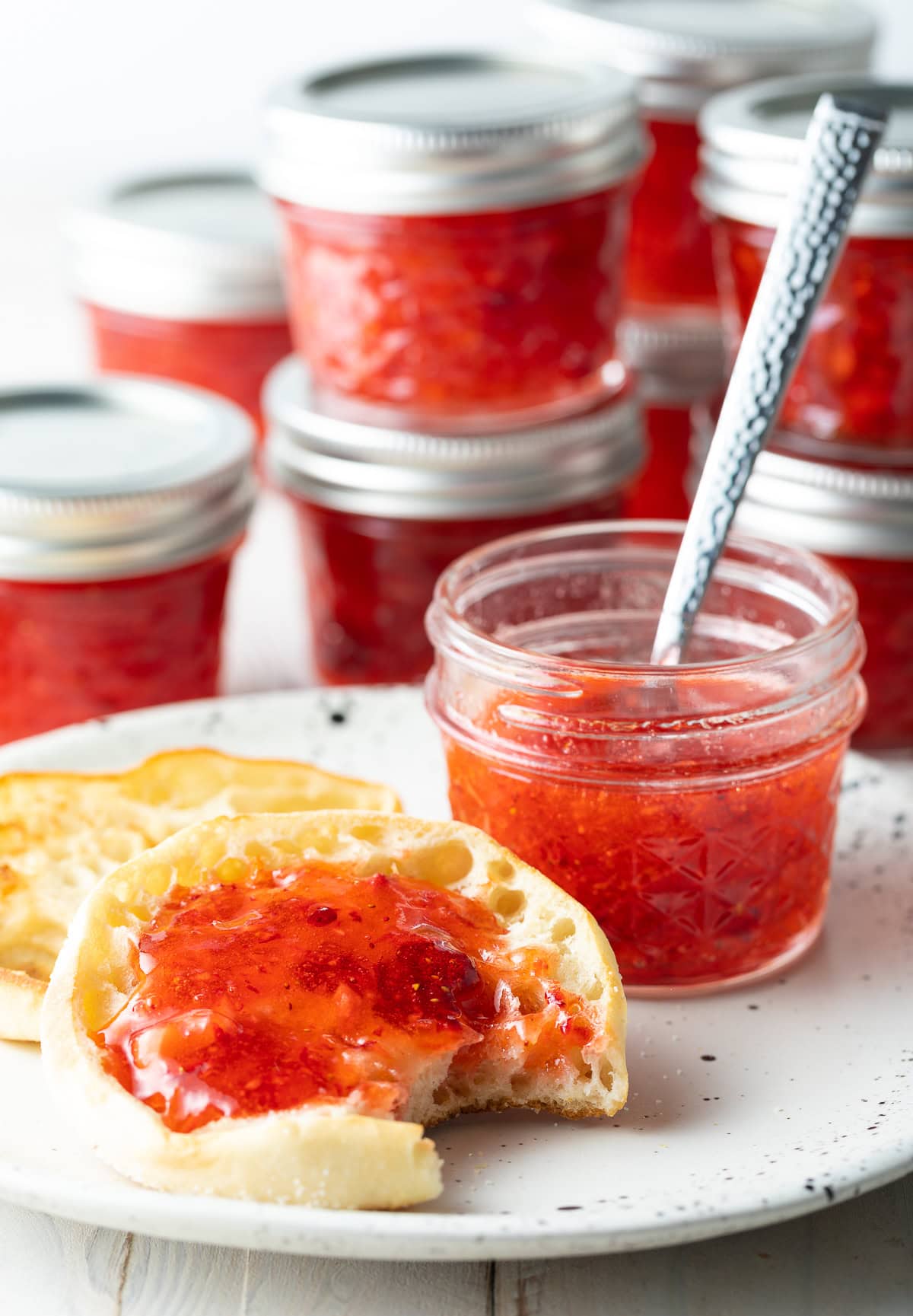 An english muffin with a bite taken out of it, strawberry freezer jam spread on top. A jar of jam is next to the muffin.