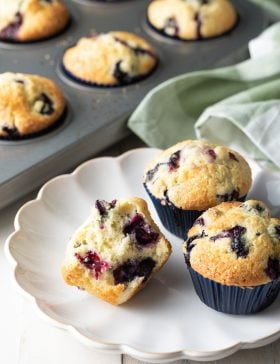 A white plate with three lemon blueberry muffins beside a baking tray of blueberry muffins.
