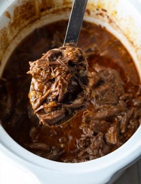 Metal spoon holding shredded roast beef from slow cooker to camera.