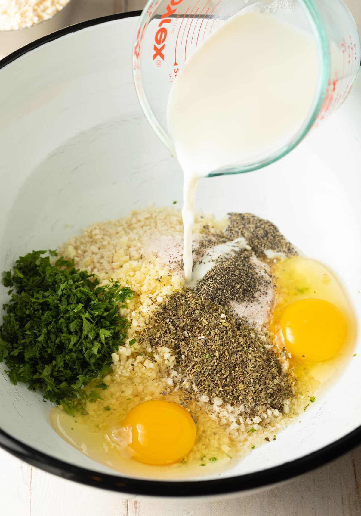 Eggs, breadcrumbs and seasonings in a mixing bowl. A jug of milk is being poured into the bowl.