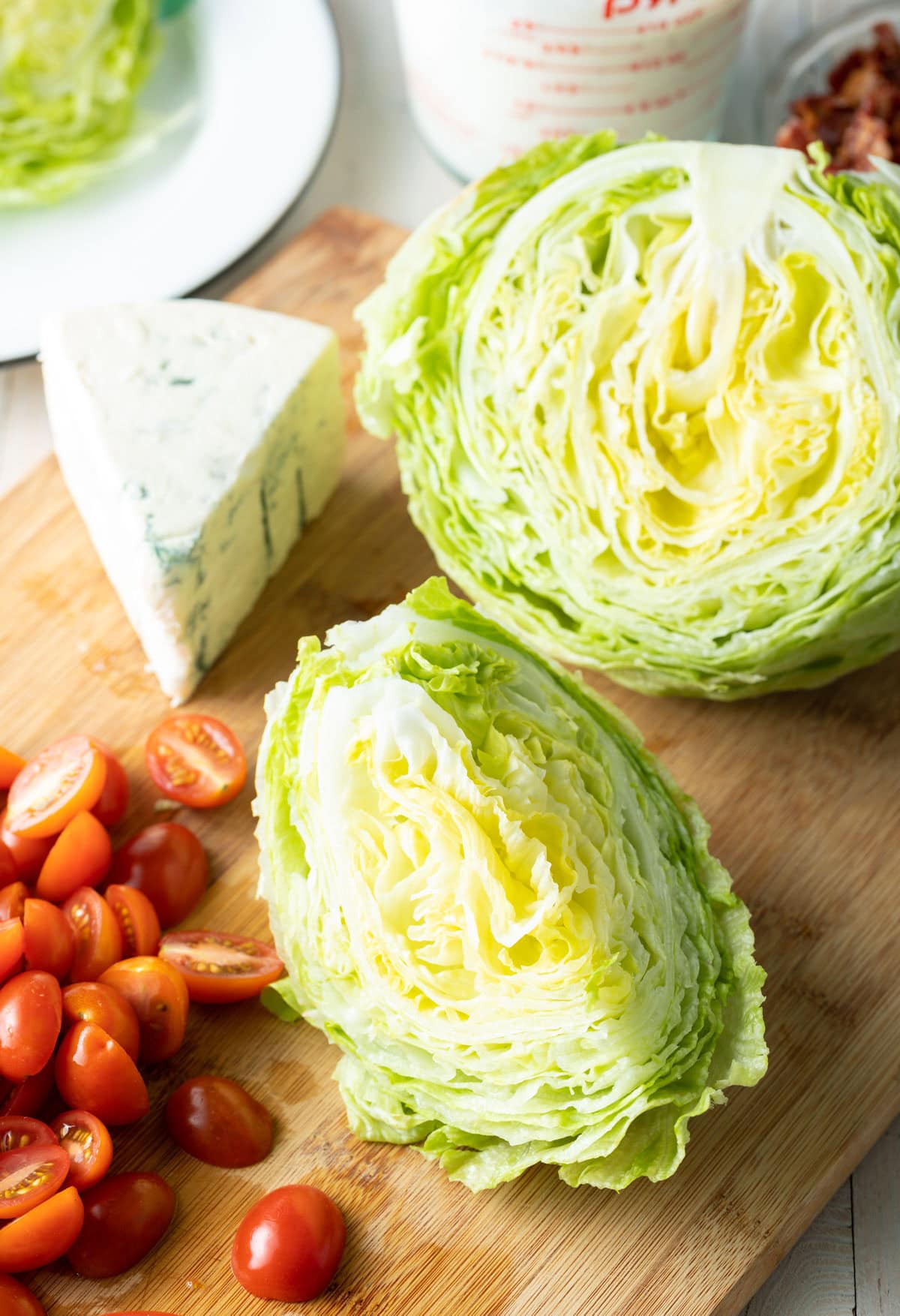 Halved and quartered iceberg, wedge of blue cheese, and pile of halved grape tomatoes on a cutting board.