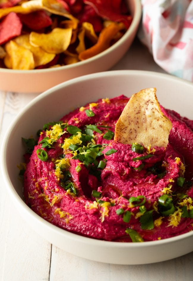 Beet dip in a white bowl topped off with sliced scallions, lemon zest and a pita chip.