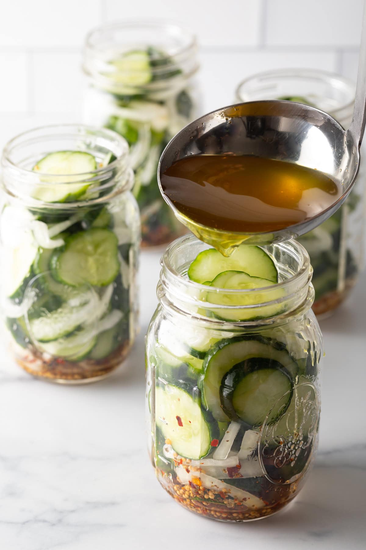 Using a metal ladle to pour pickling liquid into glass jars with sliced cucumbers and onions.