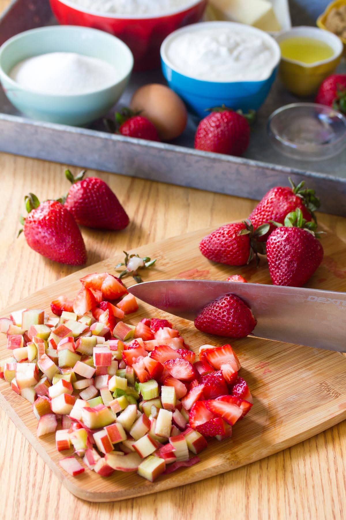 Strawberries and rhubarb being chopped on a wooden cutting board. A metal baking tray of measured ingredients for strawberry and rhubarb muffins is in the background.