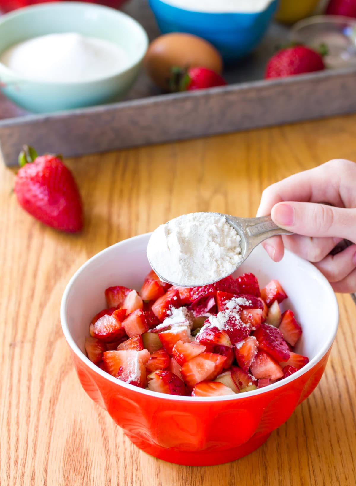 A tablespoon of flour being sprinkled over a red and white bowl of chopped strawberries and rhubarb.
