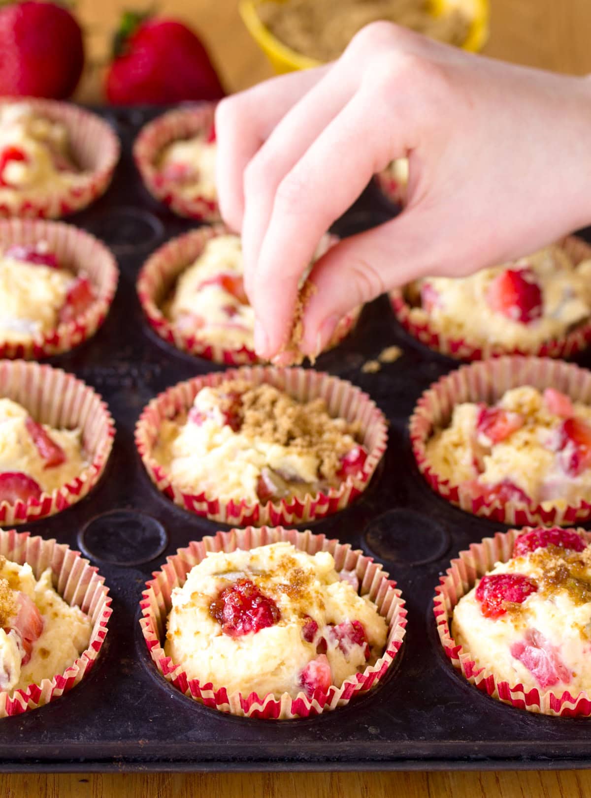 A woman's hand sprinkling brown sugar over a pan of uncooked muffins in red paper liners.