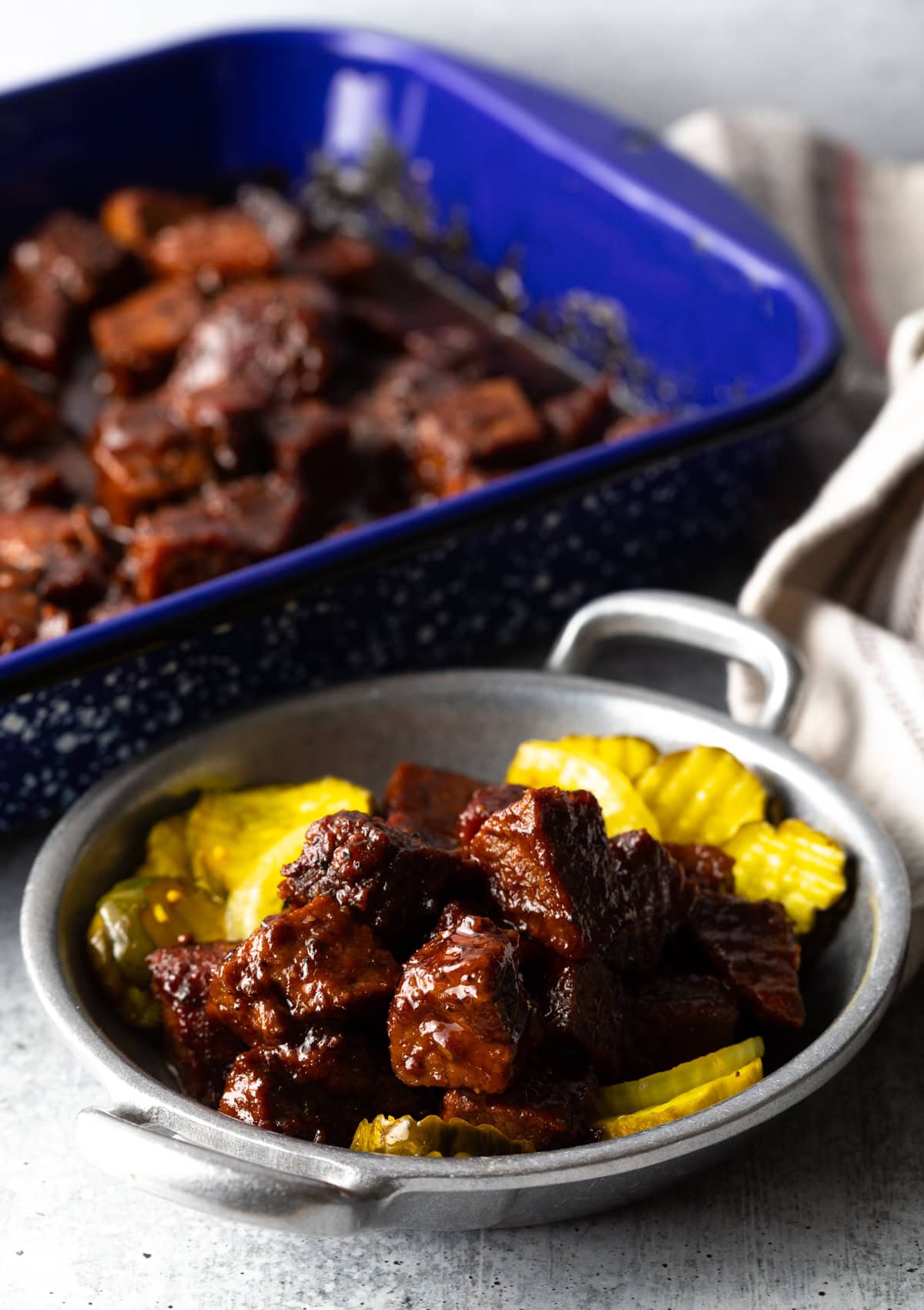 A metal dish of burnt ends and bread and butter pickles with a baking dish of more brisket burnt ends in the background.