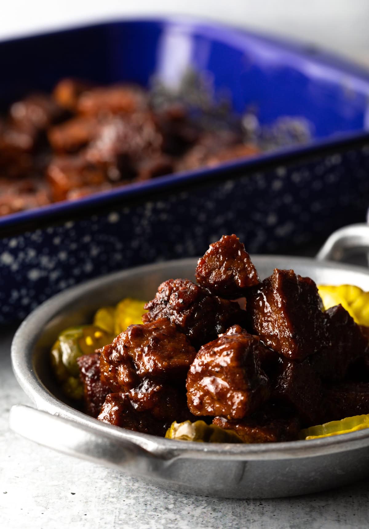Close up of a metal dish of brisket burnt ends and bread and butter pickles. There is a blue speckled baking dish of more burnt ends in the background.