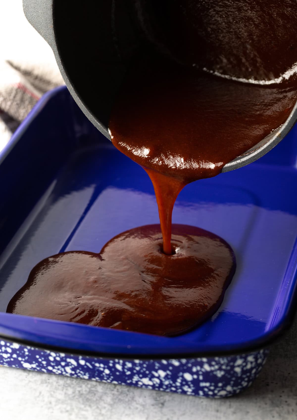 Thick and glossy barbecue sauce being poured from a cooking pot into a blue speckled roasting dish.