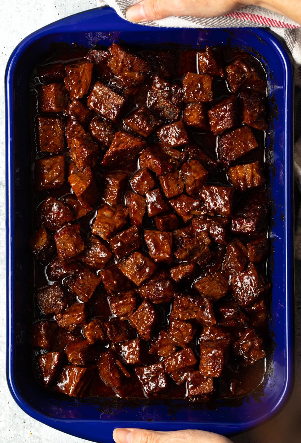 Close up of a roasting dish of burnt ends. Hands and a kitchen towel are just visible holding the dish by both ends.