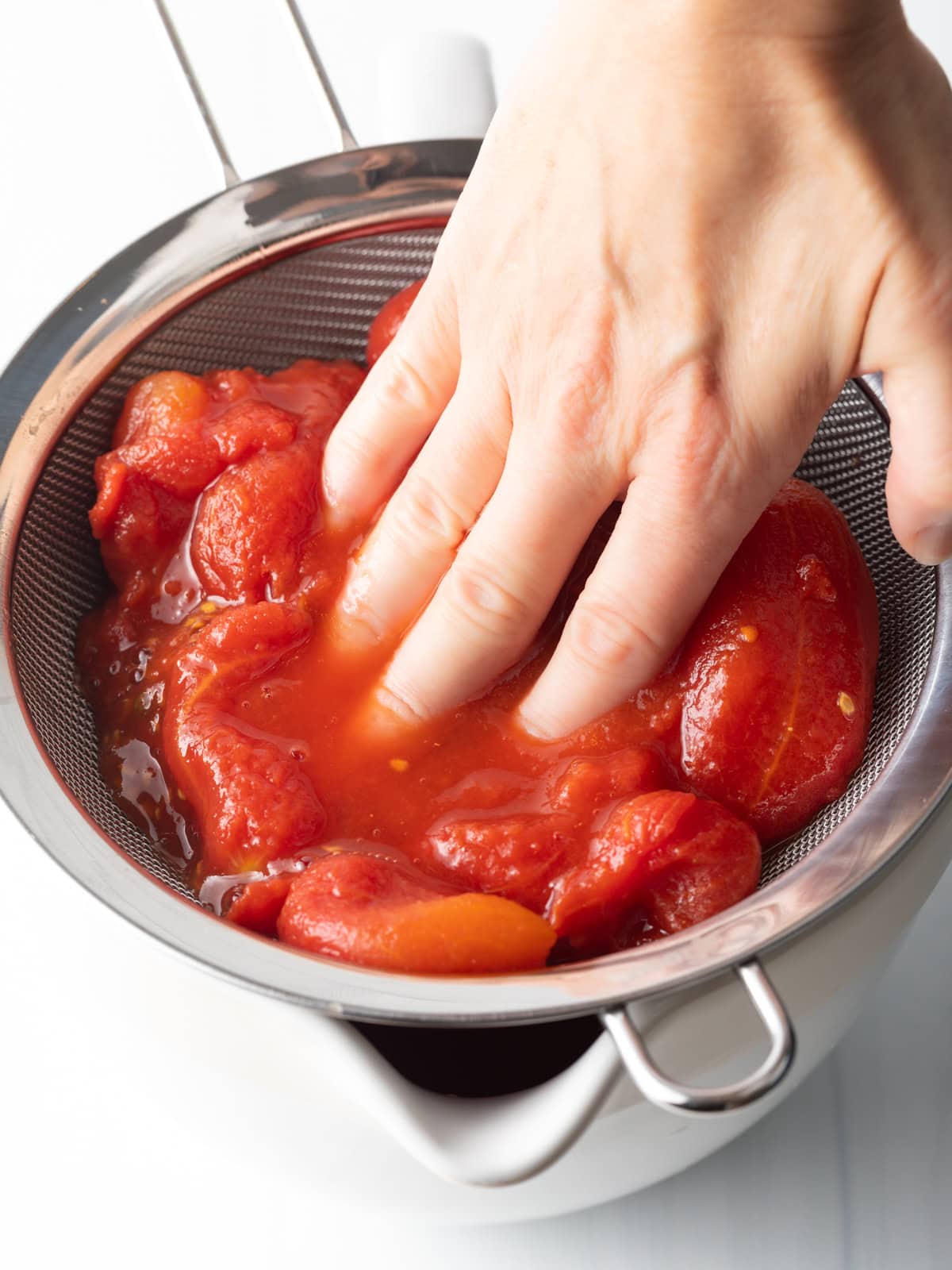 Hand smooshing whole canned tomatoes through a sieve.