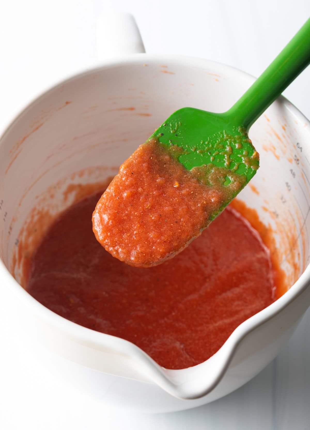 White mixing pitcher filled with tomato sauce. A green spatula is covered in the sauce and held to camera.
